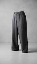 A perfectly centered shot of relaxed linen trousers in Graphite Gray, displayed against a clean concrete background. The...
